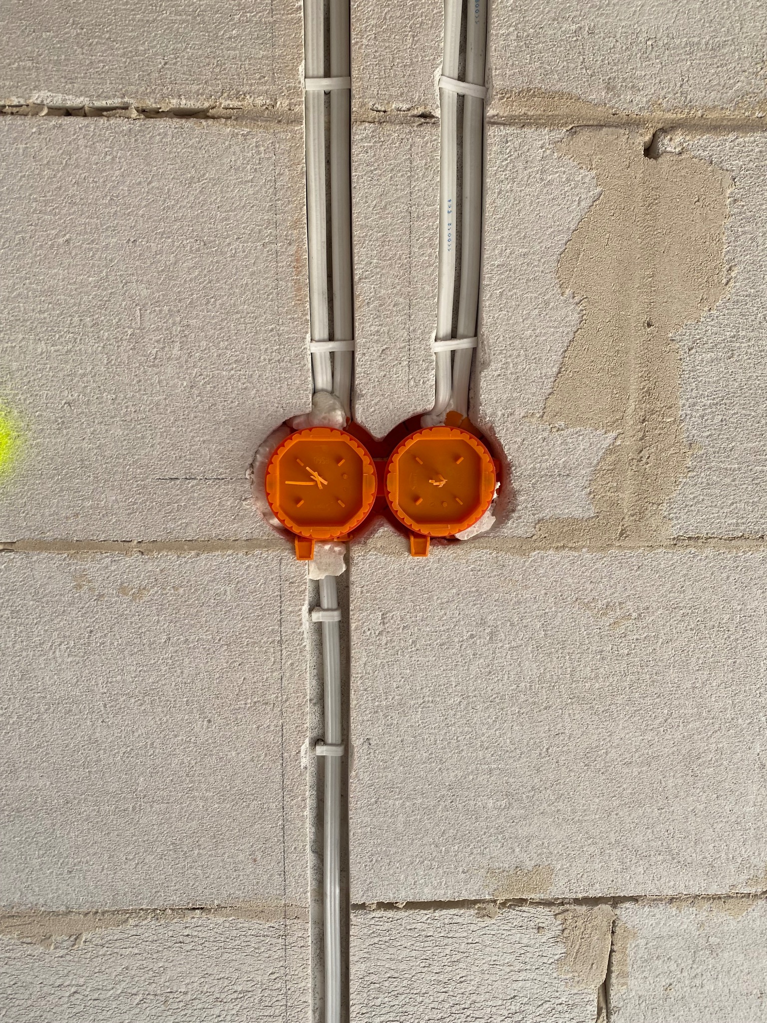 Dwa pomarańczowe gniazdka elektryczne zamontowane w ścianie z betonu komórkowego, z przymocowanymi do ściany przewodami w białych rurkach. Widoczne ślady po zaprawie.