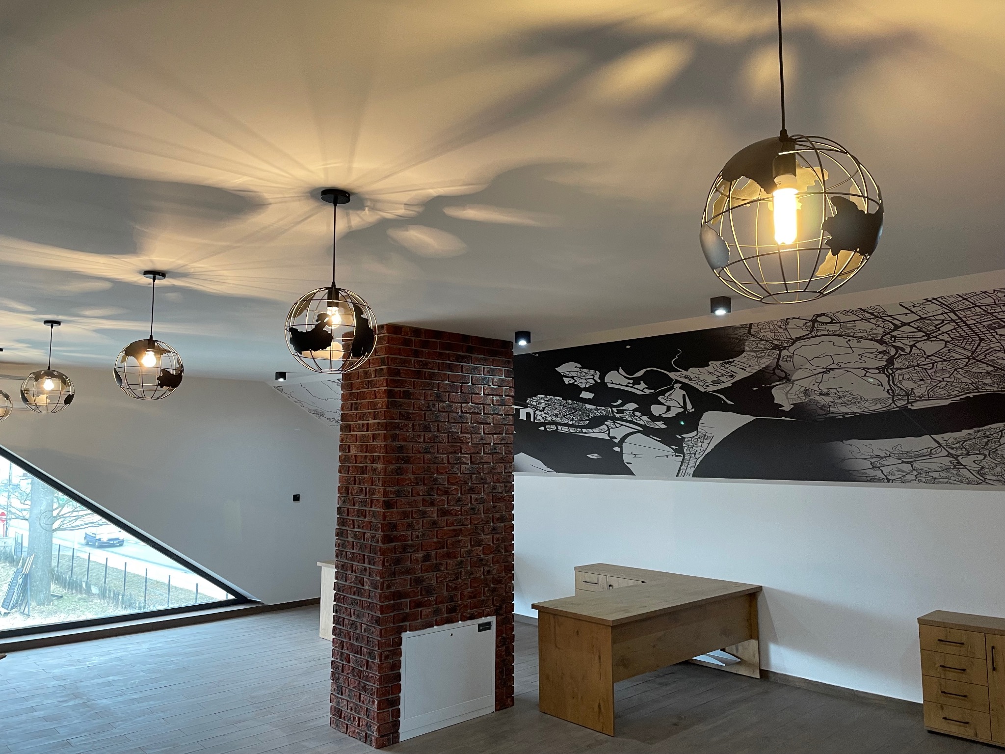 Nowoczesne wnętrze biurowe z ceglanym filarem, designerskimi lampami w kształcie globusów i minimalistycznym biurkiem. Duże okno z widokiem na zewnątrz.