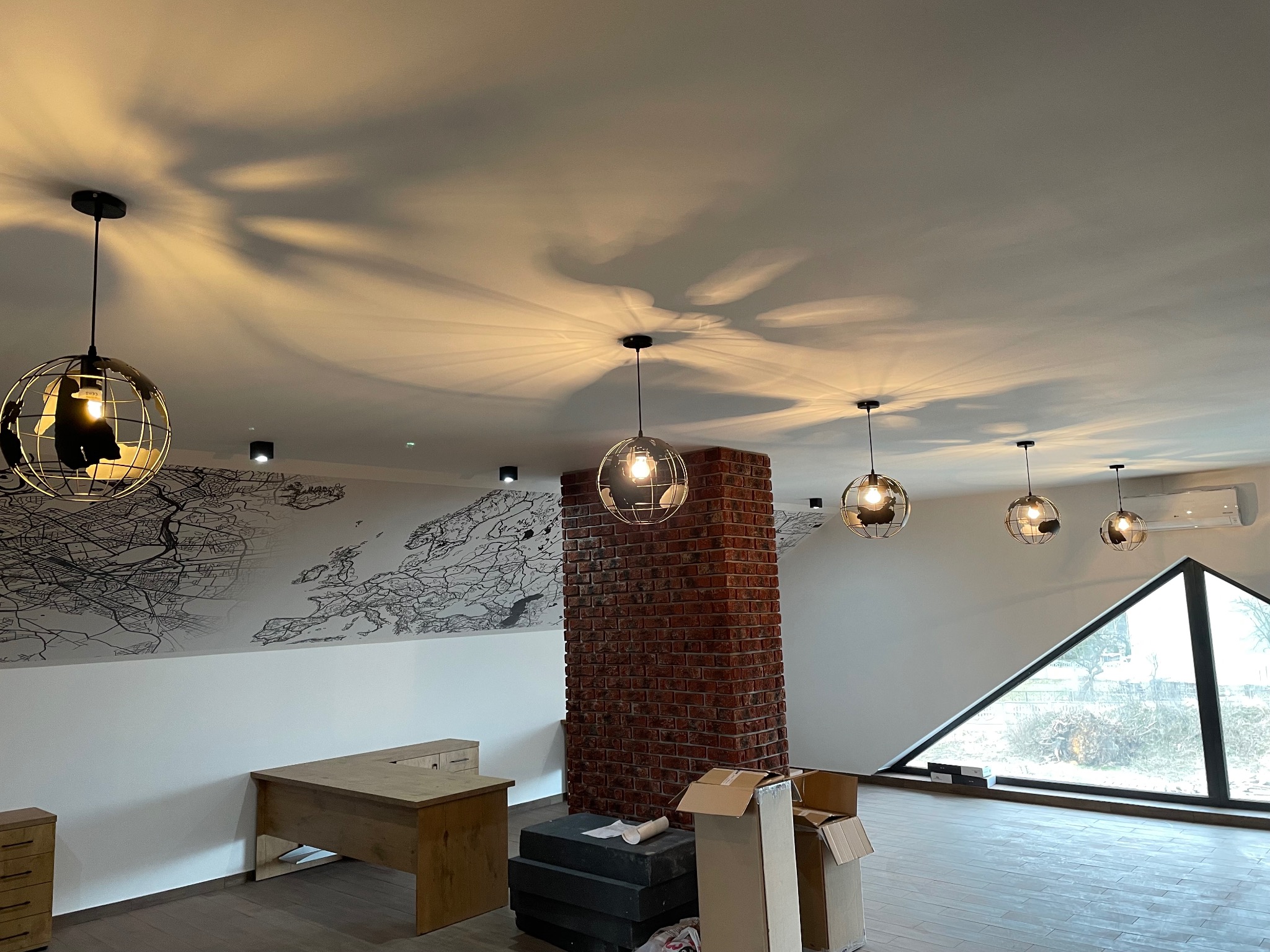 Nowoczesne wnętrze z ceglanym filarem, designerskimi lampami w kształcie globusów i mapą świata na ścianie. Widoczne biurko i okno o nietypowym kształcie.