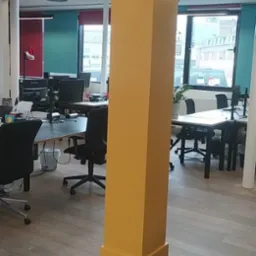 Żółta kolumna w biurze, zabezpieczona taśmą malarską u podstawy, na tle biurek i okien z widokiem na miasto.