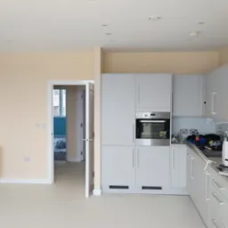 Jasne wnętrze mieszkania po remoncie, z nową zabudową kuchenną w odcieniach szarości, świeżo pomalowanymi ścianami w odcieniu kremowym, widocznym grzejnikiem i otwartymi drzwiami do kolejnego...