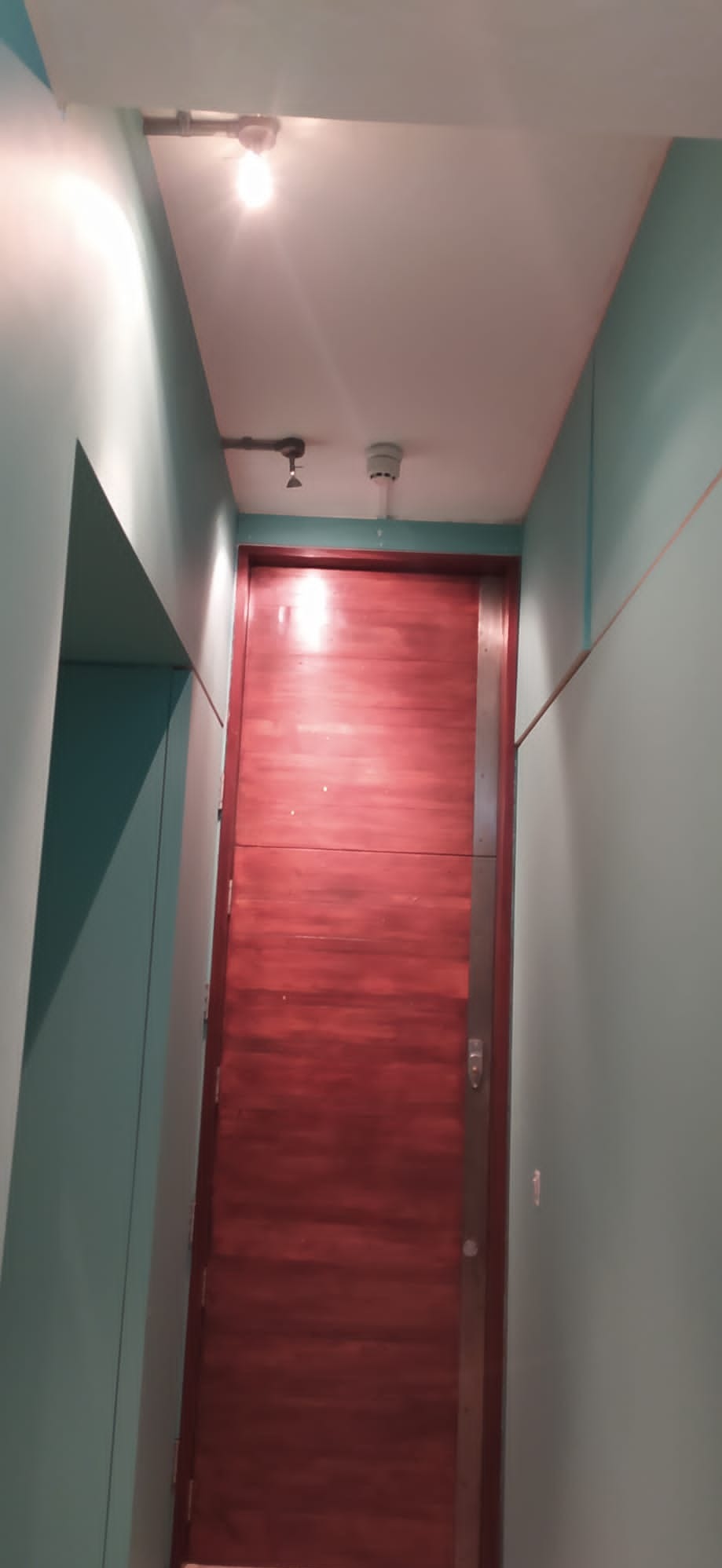 Wnętrze korytarza z pomalowanymi na turkusowo ścianami, brązowymi drzwiami z metalową listwą i widocznymi instalacjami na suficie.