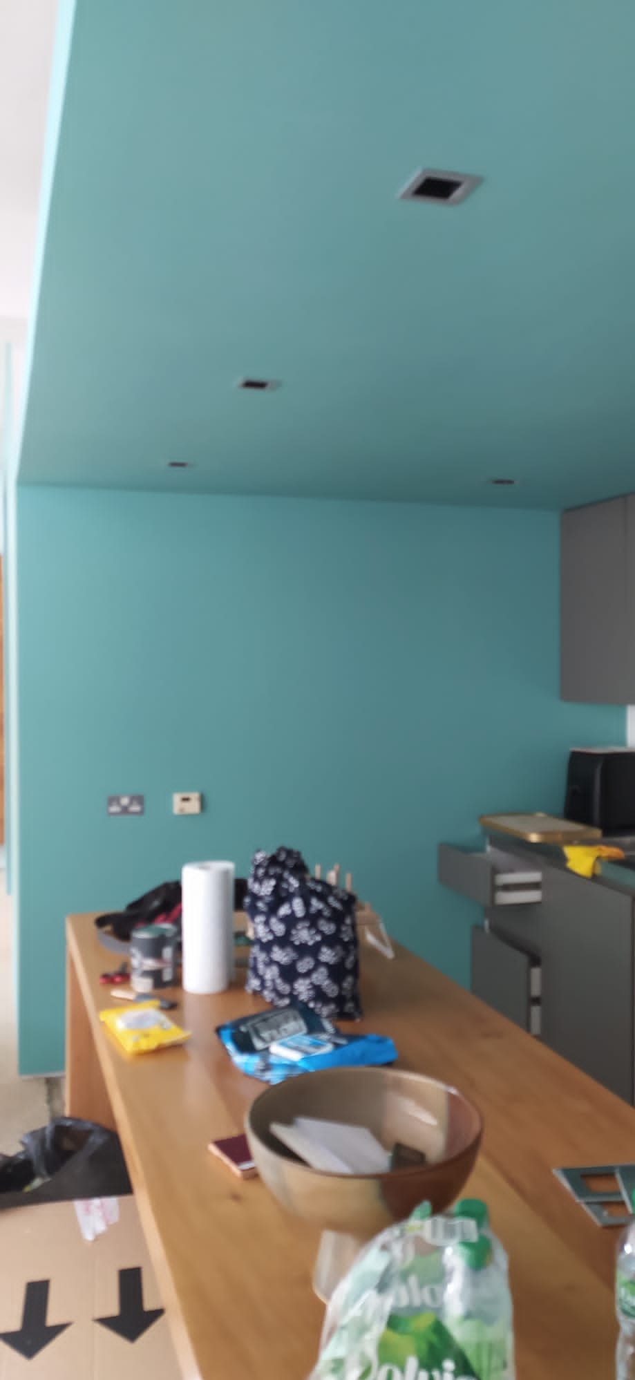 Świeżo pomalowane na turkusowo ściany i sufit w pokoju, z widocznymi gniazdkami elektrycznymi i elementami wyposażenia kuchni w tle, oraz z akcesoriami malarskimi i osobistymi leżącymi...