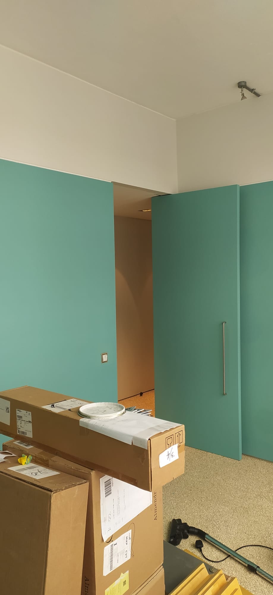 Świeżo pomalowane na turkusowo ściany z ukrytymi drzwiami przesuwnymi z długim, pionowym uchwytem, widoczne kartony i akcesoria malarskie na podłodze.