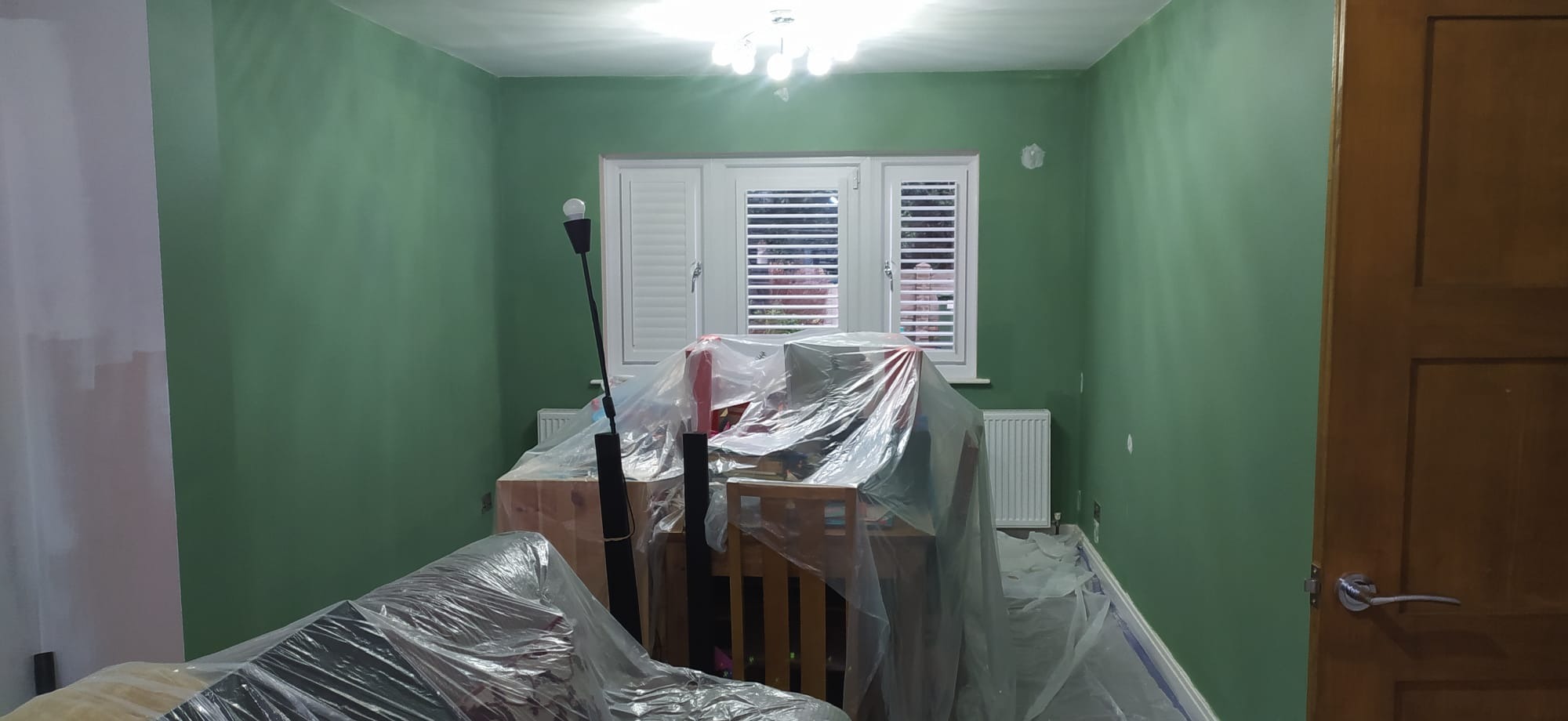 Pokój w trakcie malowania na zielono, meble zabezpieczone folią, widoczne niedomalowane fragmenty ściany przy suficie, białe okno z żaluzjami.