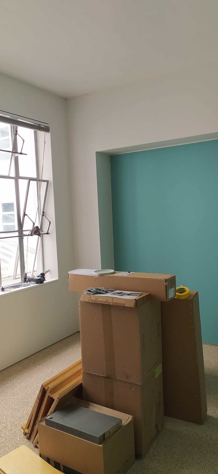 Pokój przed remontem: kartony z materiałami budowlanymi, niebieska ściana w tle, okno z widocznymi narzędziami.