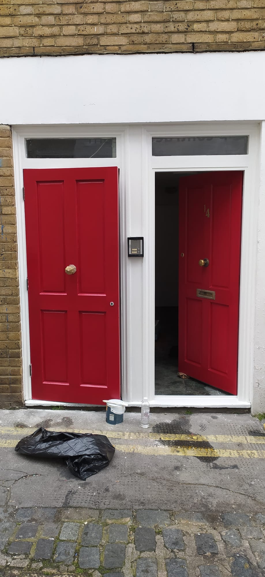 Świeżo pomalowane, jaskrawoczerwone drzwi wejściowe z białymi futrynami, z widoczną puszką farby i butelką wody na chodniku oraz czarnym workiem na śmieci.