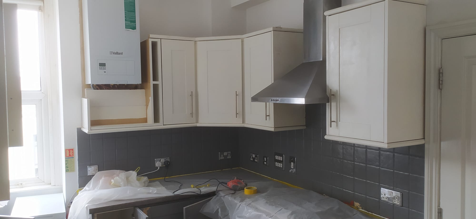 Kuchnia w trakcie remontu: białe szafki wiszące, szare płytki na ścianie, zabezpieczone blaty folią malarską, widoczny bojler i okap.