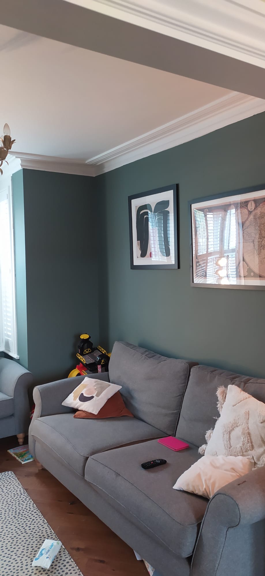 Ściana pomalowana na ciemnozielony kolor z białym, ozdobnym gzymsem pod sufitem, szara kanapa z poduszkami, na ścianie wiszą dwa obrazy w czarnych ramkach.