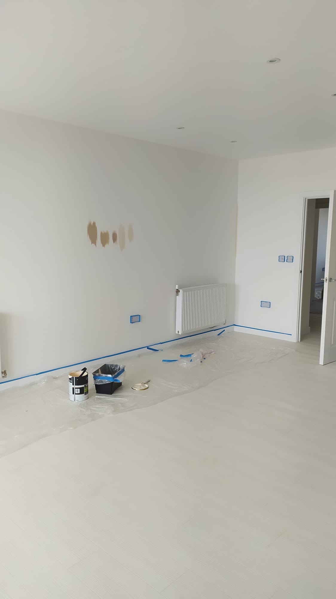 Przygotowania do malowania pokoju: zabezpieczona folią podłoga, taśmą malarską oklejone gniazdka i grzejnik, puszka z farbą, kuweta i pędzel przy ścianie z plamami po testowaniu koloru.