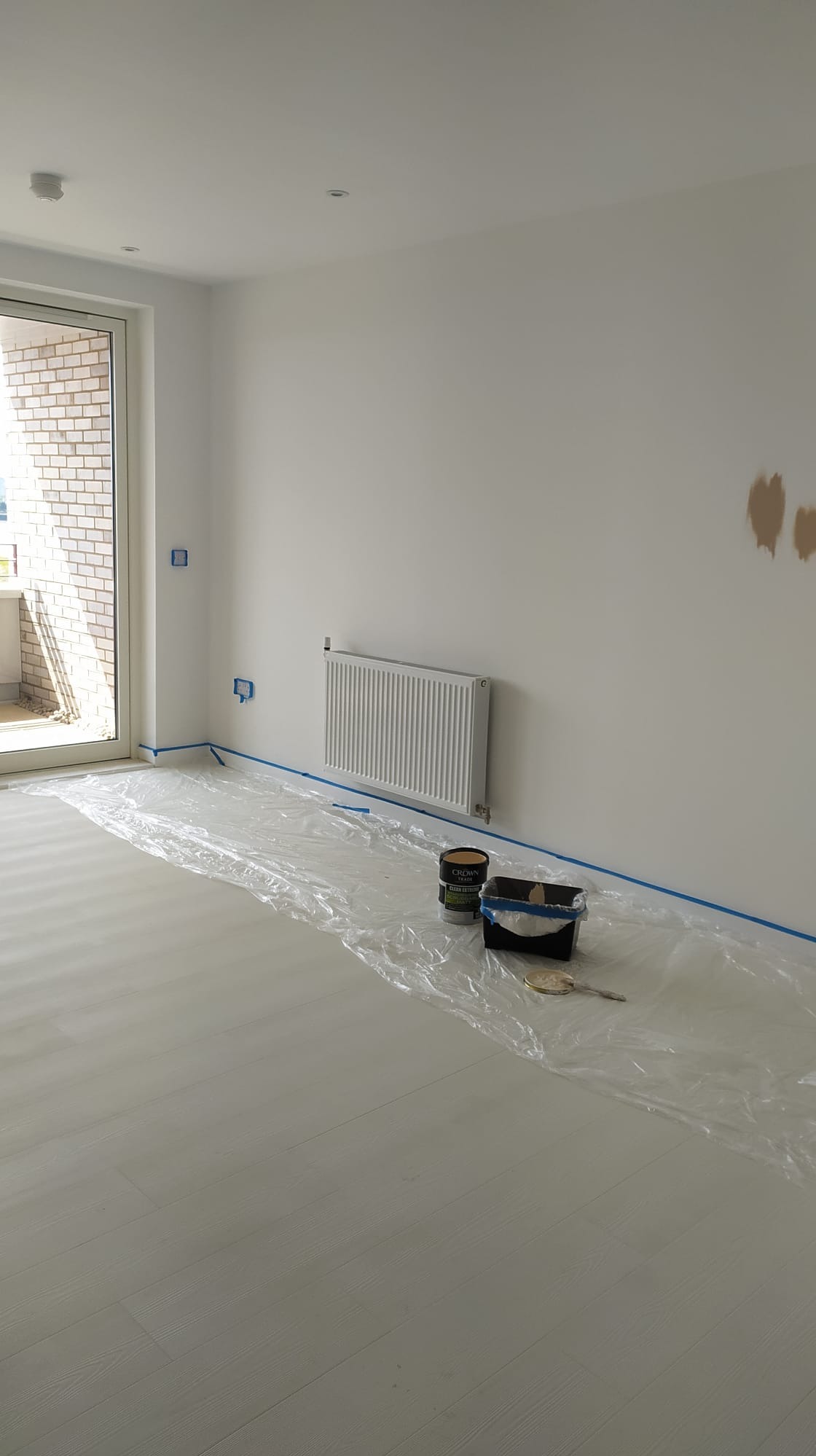 Przygotowanie do malowania pokoju: zabezpieczona folią podłoga z panelami, grzejnik i narożniki oklejone taśmą malarską, widoczne puszki z farbą i pędzel.