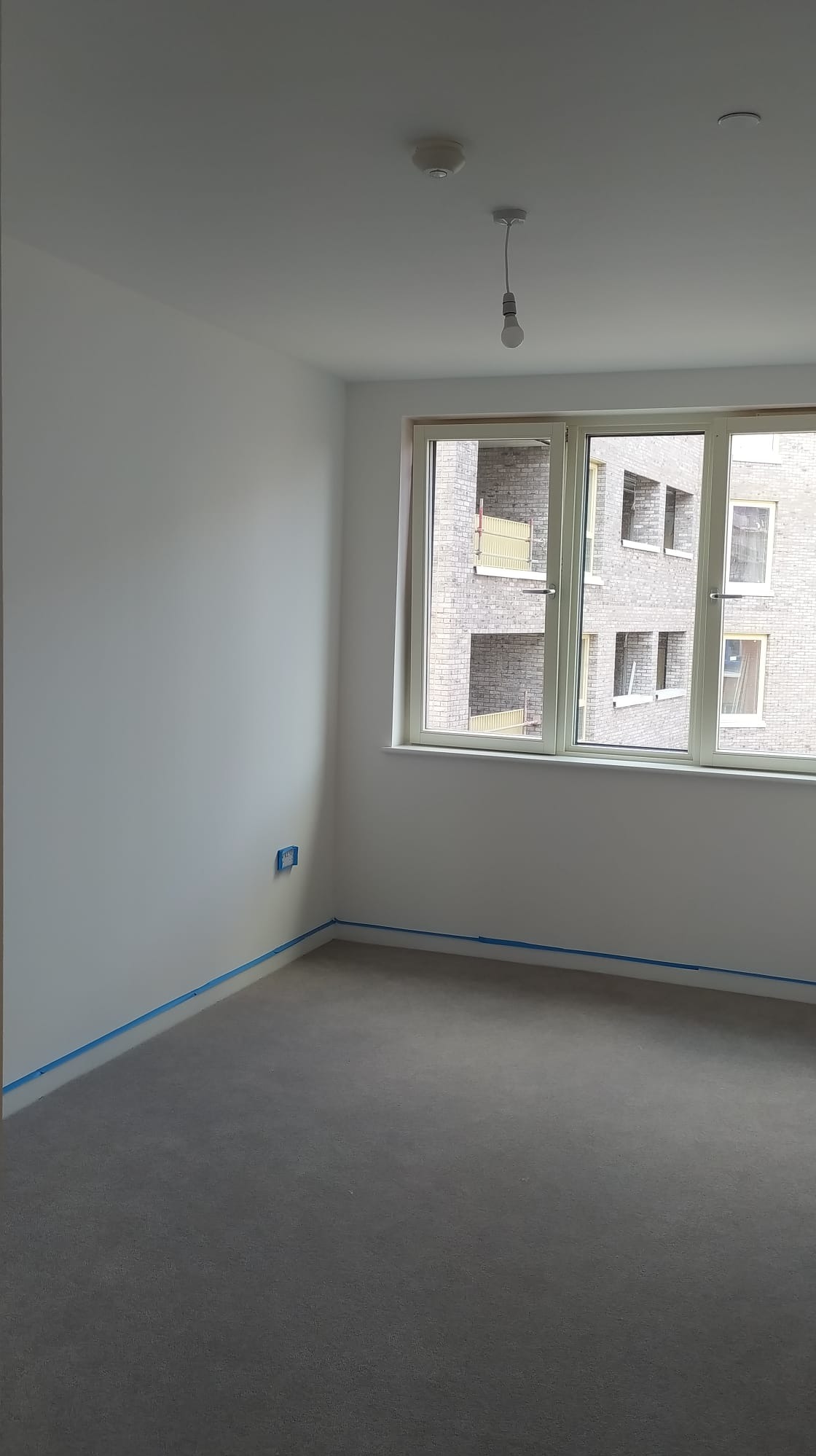Pokój przygotowany do malowania: ściany zabezpieczone taśmą malarską, szary dywan, widok z okna na budynek z cegły.