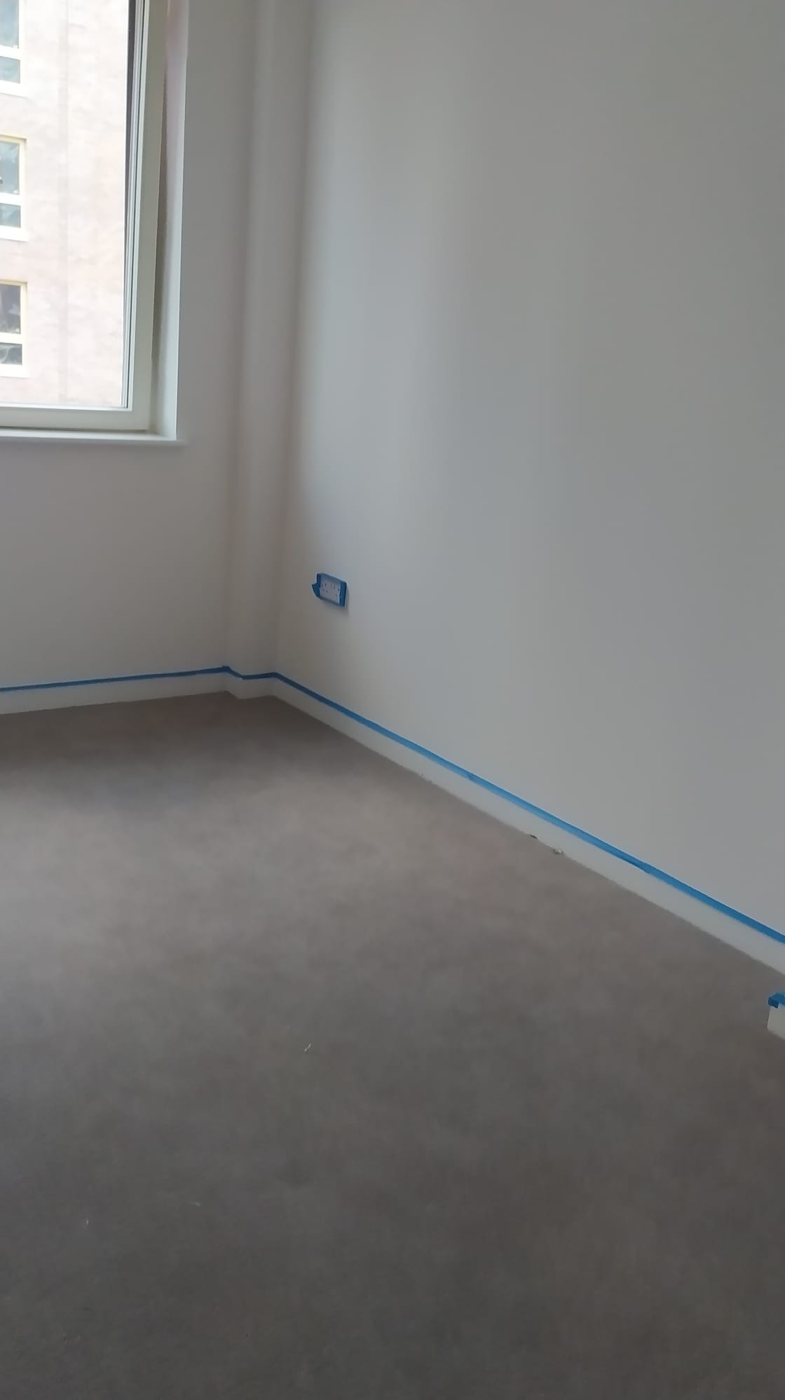 Przygotowanie do malowania: Pokój z zabezpieczonymi listwami przypodłogowymi i gniazdkiem za pomocą niebieskiej taśmy malarskiej, gotowy do nałożenia farby.