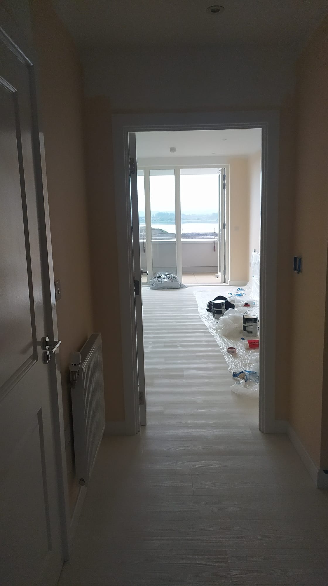 Korytarz w trakcie przygotowań do malowania, podłoga zabezpieczona folią malarską, widoczne puszki z farbą i narzędzia malarskie, w tle przeszklone drzwi balkonowe z widokiem na wodę.