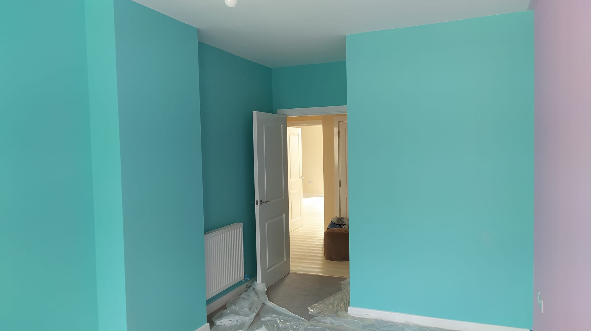Świeżo pomalowane na turkusowo wnętrze pokoju z otwartymi białymi drzwiami, widoczna grzejnik i folia ochronna na podłodze.