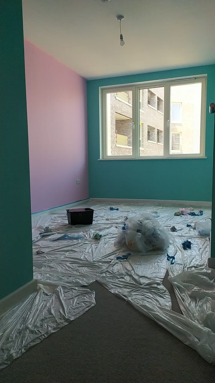 Pokój w trakcie malowania: ściany w kolorach turkusowym i różowym, podłoga zabezpieczona folią malarską, widoczne wiaderko i worki z materiałami.
