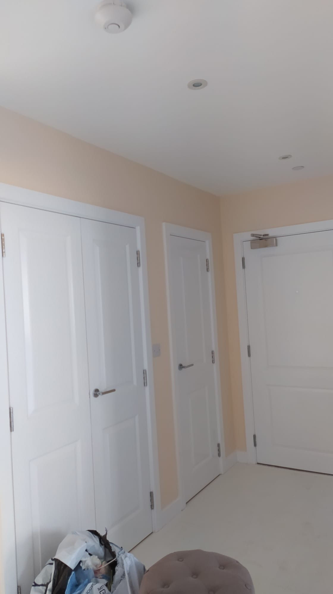 Jasne wnętrze po remoncie, ściany pomalowane na delikatny odcień brzoskwini, trzy białe drzwi z srebrnymi klamkami, widoczny fragment podłogi i worki z odpadami budowlanymi.