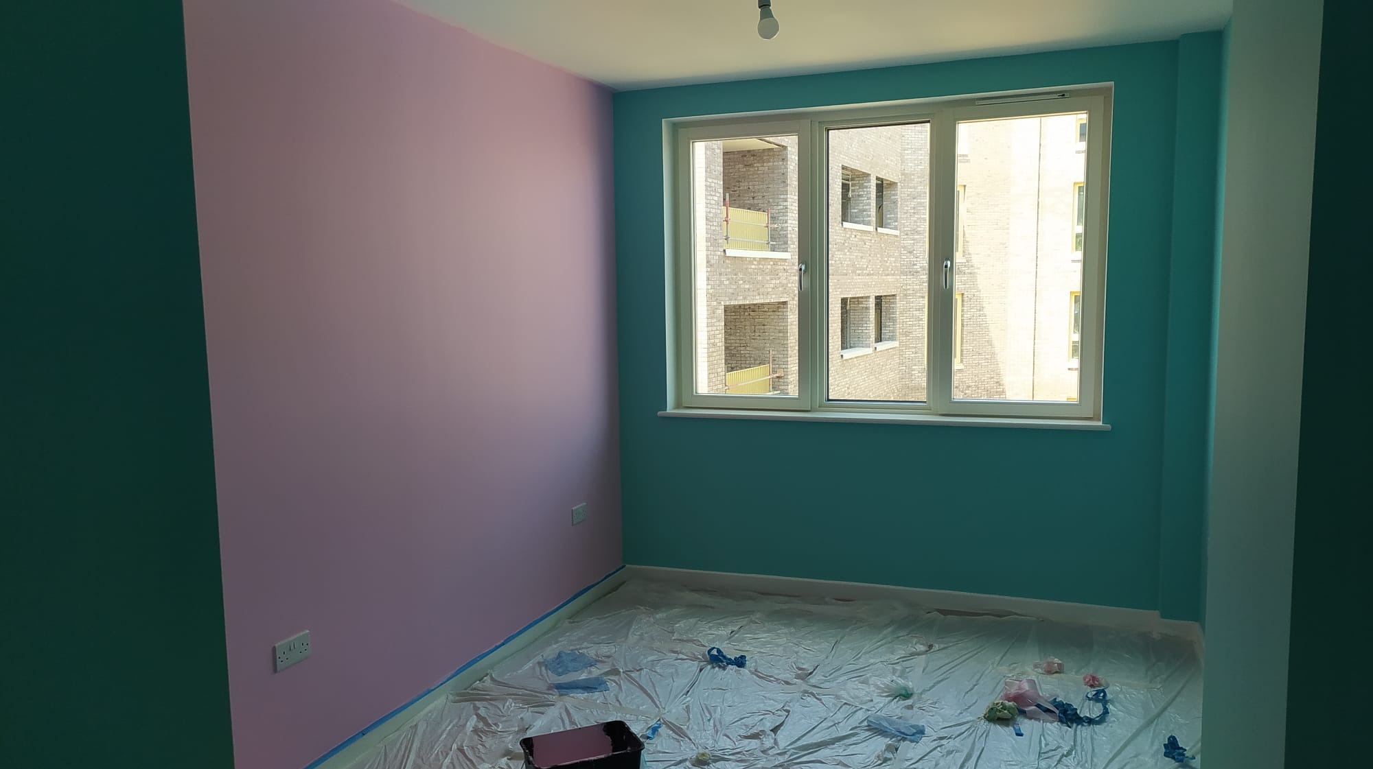 Pokój po malowaniu ścian na różowo, turkusowo i zielono, zabezpieczona folią podłoga, widok z okna na budynek z cegły.