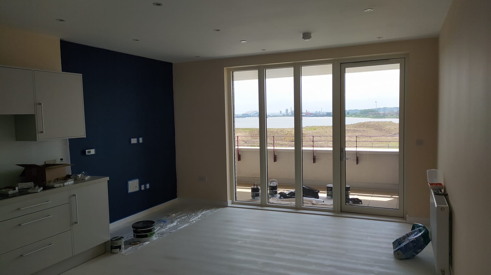 Wnętrze mieszkania w trakcie prac: ściana pomalowana na granatowo, reszta ścian w odcieniach beżu, widoczne puszki z farbą i folia ochronna na podłodze, okno balkonowe z widokiem na morze.