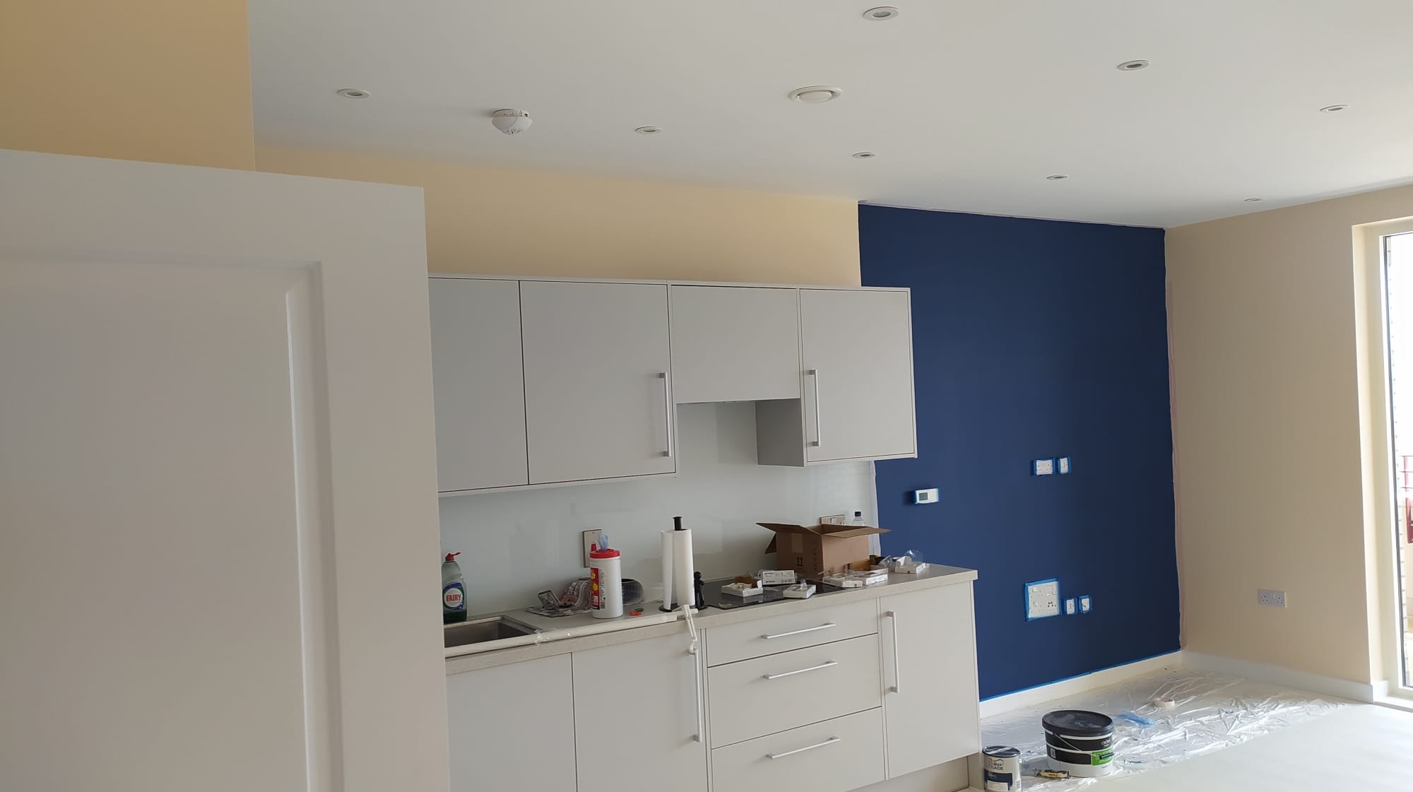 Wnętrze mieszkania w trakcie malowania: ściana pomalowana na granatowo, druga ściana w kolorze kremowym, białe meble kuchenne, zabezpieczona folią podłoga, puszki z farbą przy ścianie.