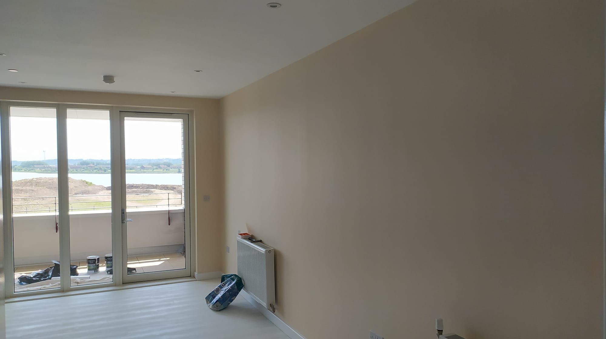 Jasne, świeżo pomalowane wnętrze z widokiem na jezioro przez przesuwne drzwi balkonowe; puszki z farbą i osłonięty grzejnik stoją przy ścianie.