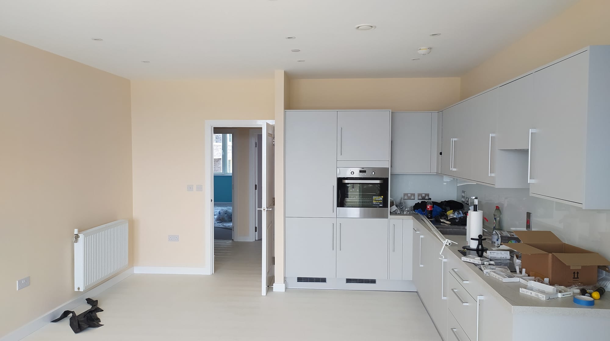 Jasne wnętrze mieszkania po remoncie, z nową zabudową kuchenną w odcieniach szarości, świeżo pomalowanymi ścianami w odcieniu kremowym, widocznym grzejnikiem i otwartymi drzwiami do kolejnego...