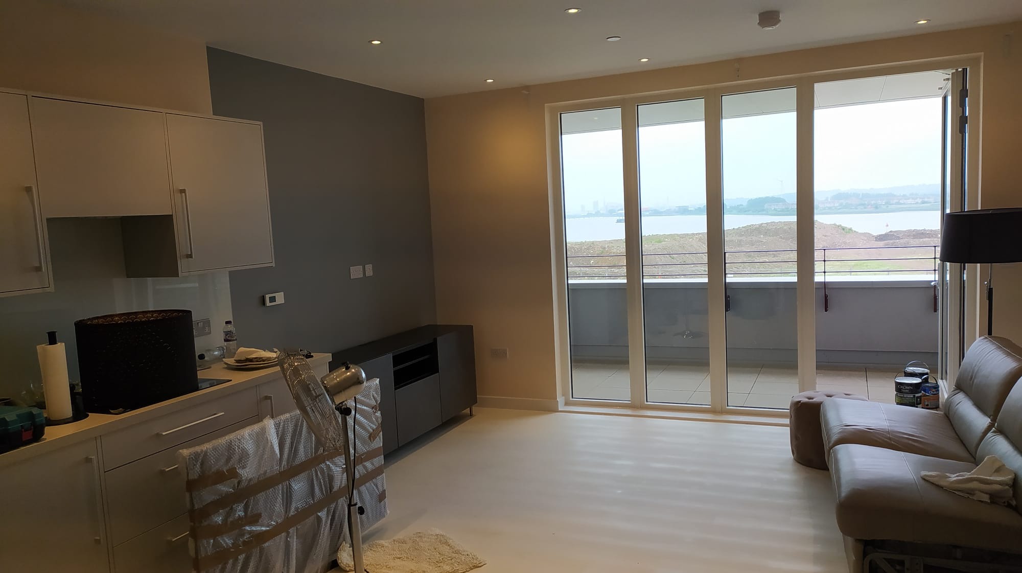 Wnętrze mieszkania po remoncie: świeżo pomalowane ściany w odcieniach szarości i beżu, białe meble kuchenne i szafka RTV, duże okno balkonowe z widokiem na wodę, częściowo zakryte meble folią...