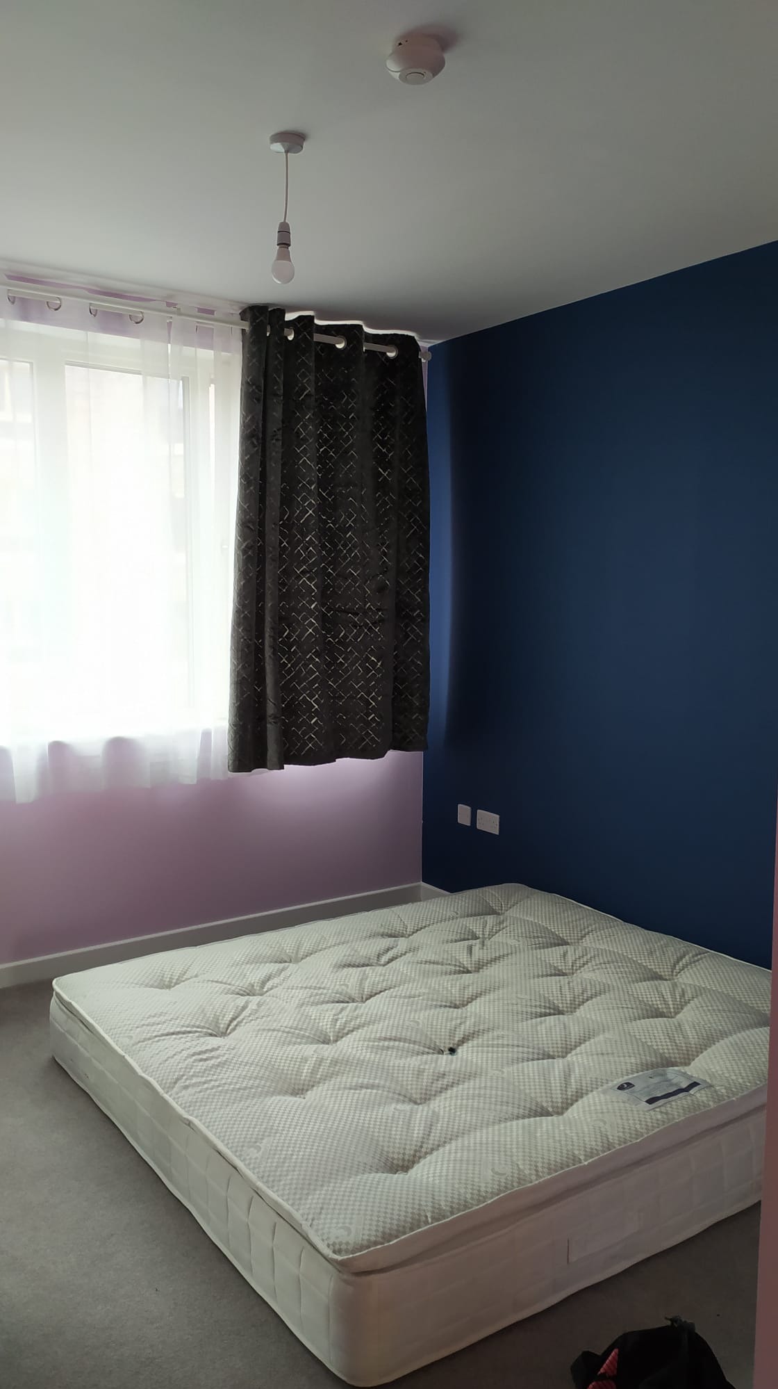 Pokój po malowaniu: ściana pomalowana na granatowo sąsiaduje z fioletową ścianą, na podłodze materac bez ramy łóżka, zasłony w oknie