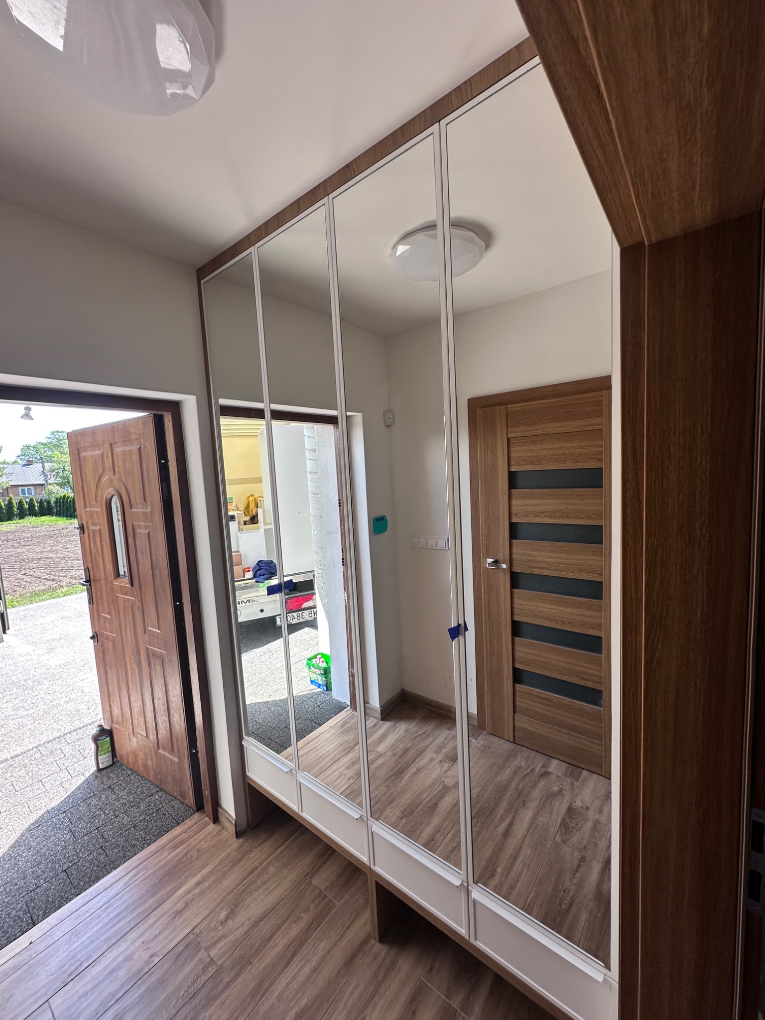 Nowoczesna szafa wnękowa z lustrzanymi drzwiami przesuwnymi, wkomponowana w przedpokój z drewnianą podłogą i drzwiami. Widok na otwarte drzwi wejściowe.