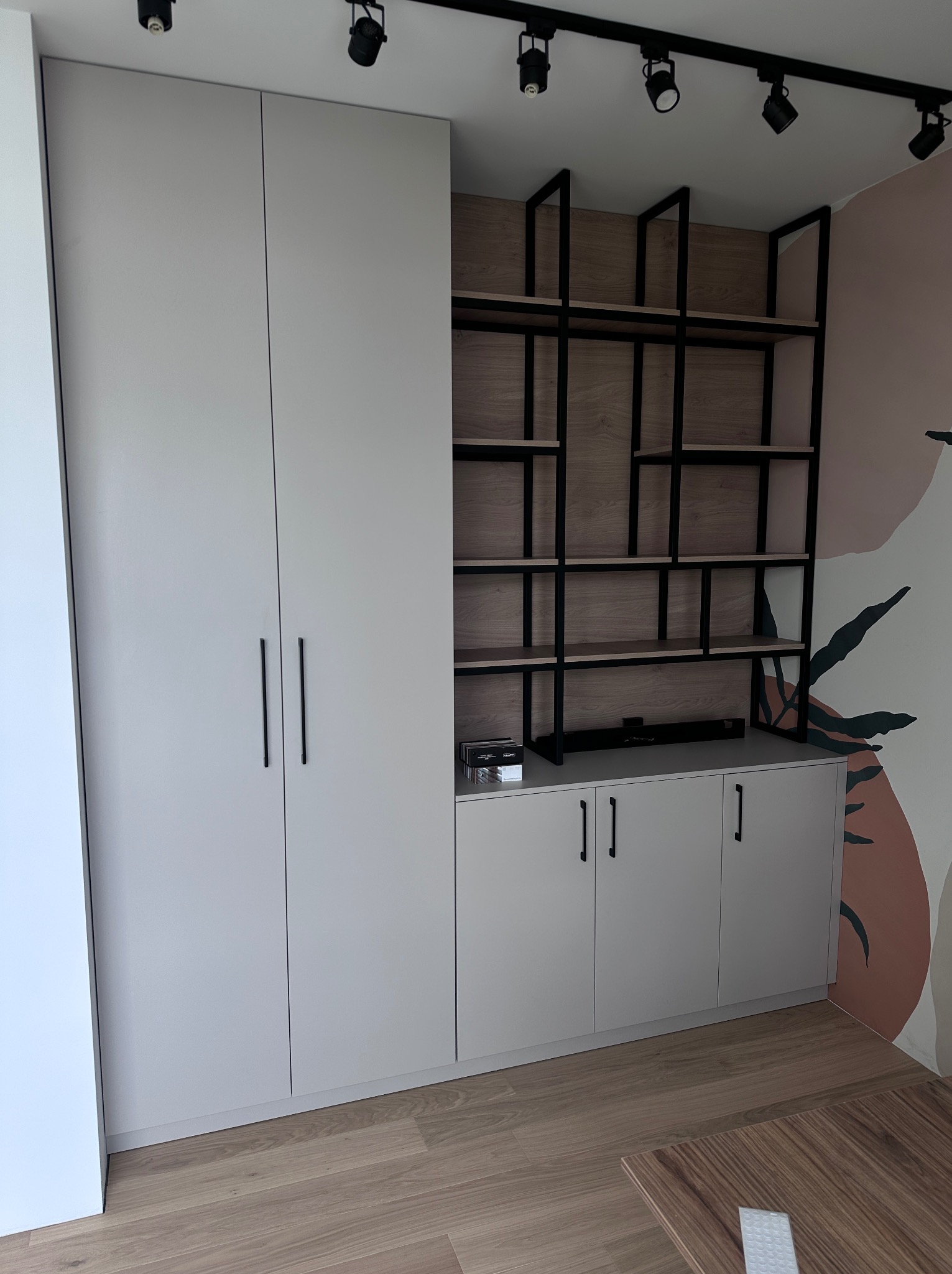 Biało-szara szafa wnękowa z czarnymi uchwytami i regałem z czarną metalową konstrukcją oraz drewnianymi półkami, na tle ściany z abstrakcyjnym wzorem.