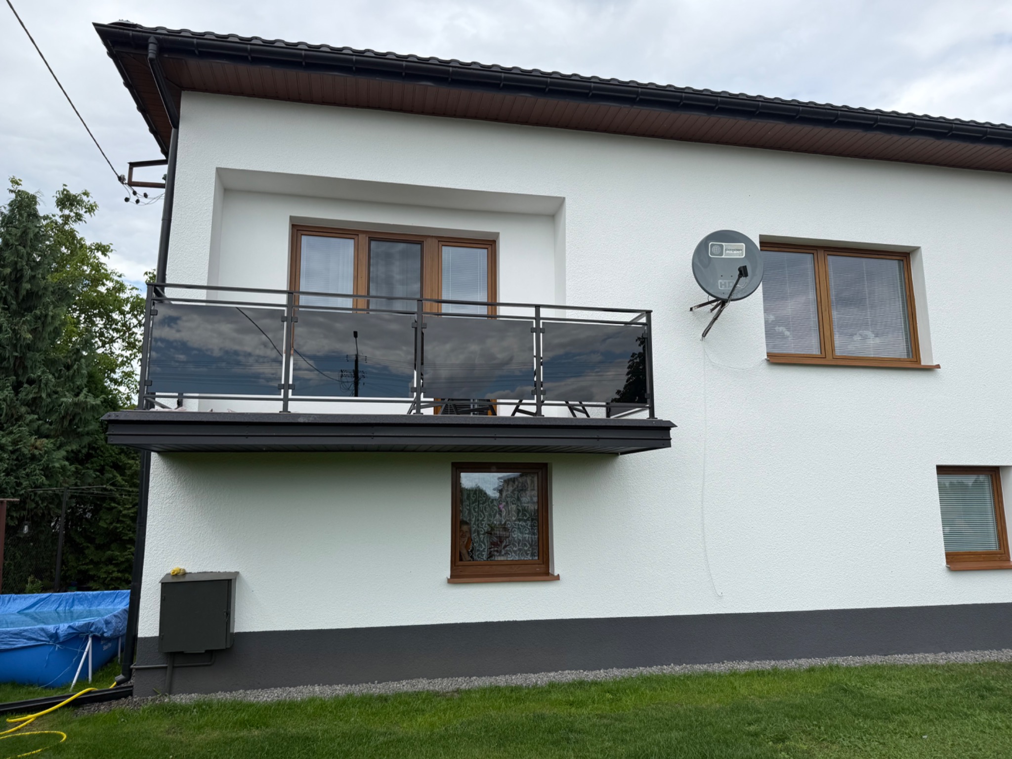 Nowoczesna balustrada balkonowa z czarnymi, metalowymi ramami i szklanymi panelami, widoczna na tle białej elewacji budynku z brązowymi oknami.