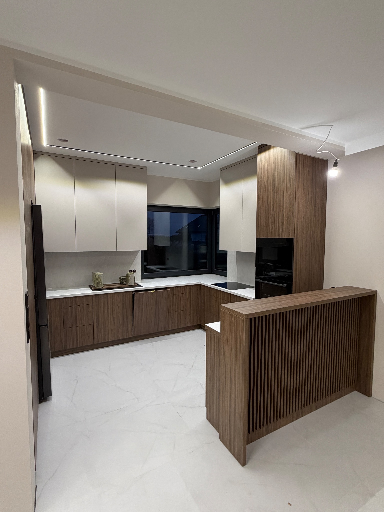 Nowoczesna kuchnia z drewnianymi i białymi szafkami, marmurowym blatem i oświetleniem LED. Widoczny fragment wyspy kuchennej z dekoracyjnymi listwami.