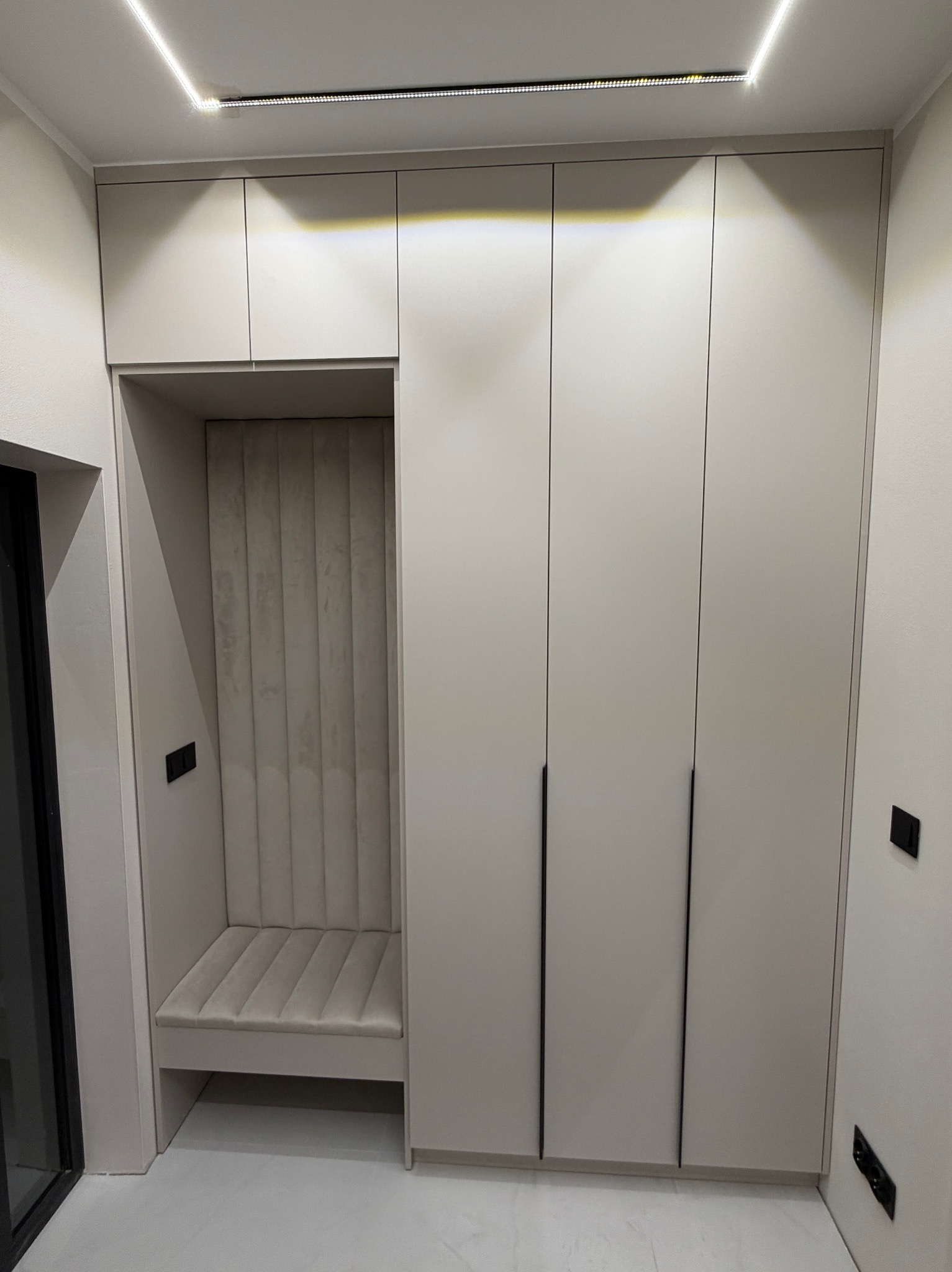 Jasna szafa wnękowa z siedziskiem i tapicerowanym panelem ściennym, minimalistyczne czarne uchwyty, oświetlenie LED na suficie, gładkie fronty.