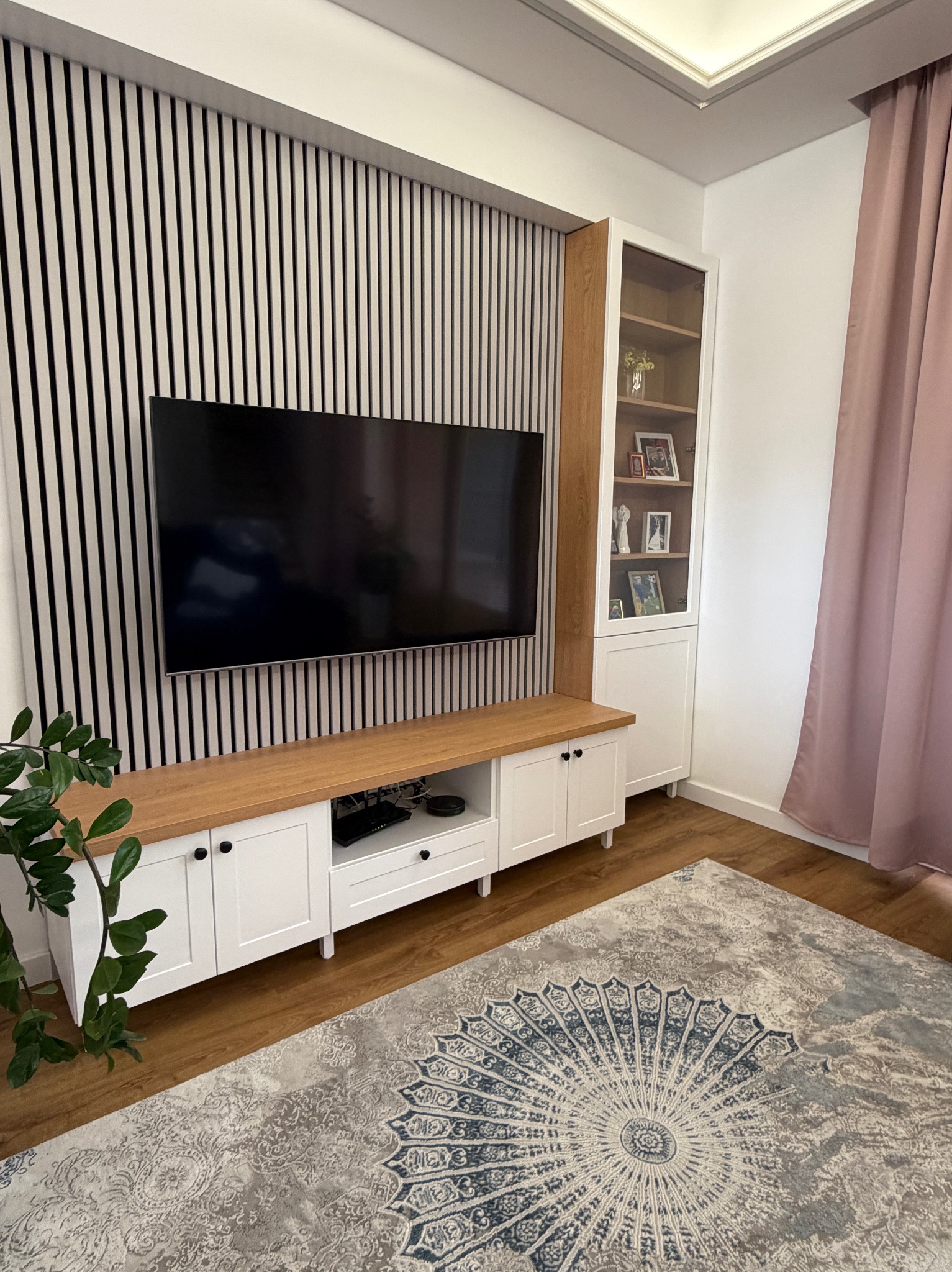 Biała szafka RTV z drewnianym blatem pod telewizorem na tle ściany z pionowych lameli. Obok witryna z przeszklonymi drzwiami i dekoracyjny dywan.