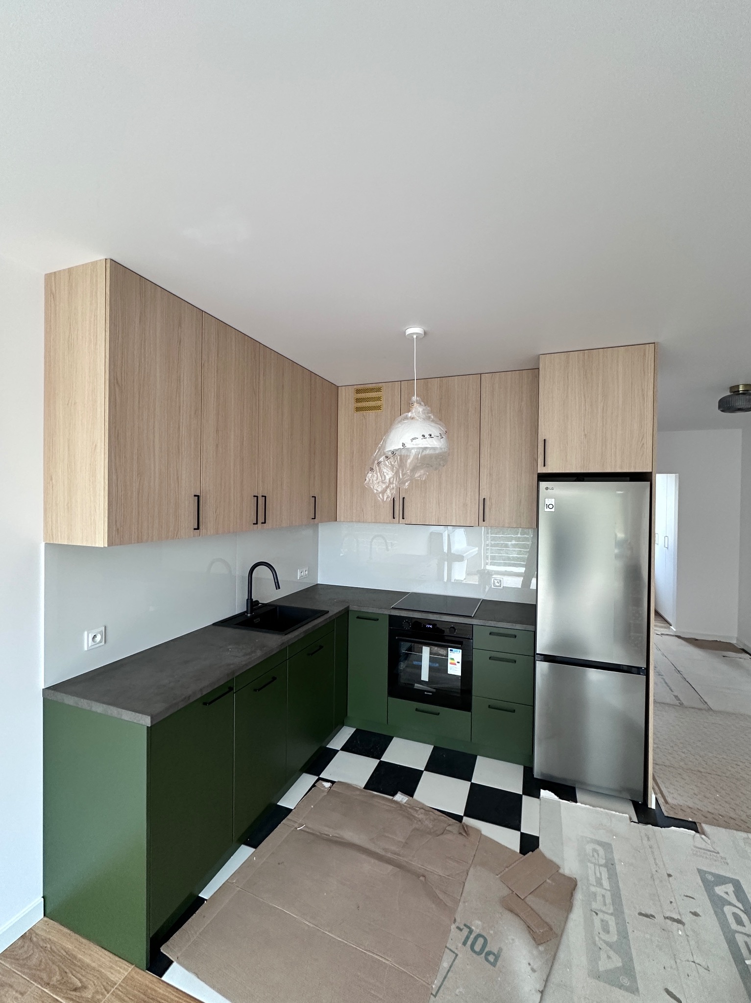 Nowoczesna kuchnia z zielonymi szafkami dolnymi i drewnopodobnymi górnymi, czarno-biała podłoga, srebrna lodówka. Lampa w folii ochronnej.