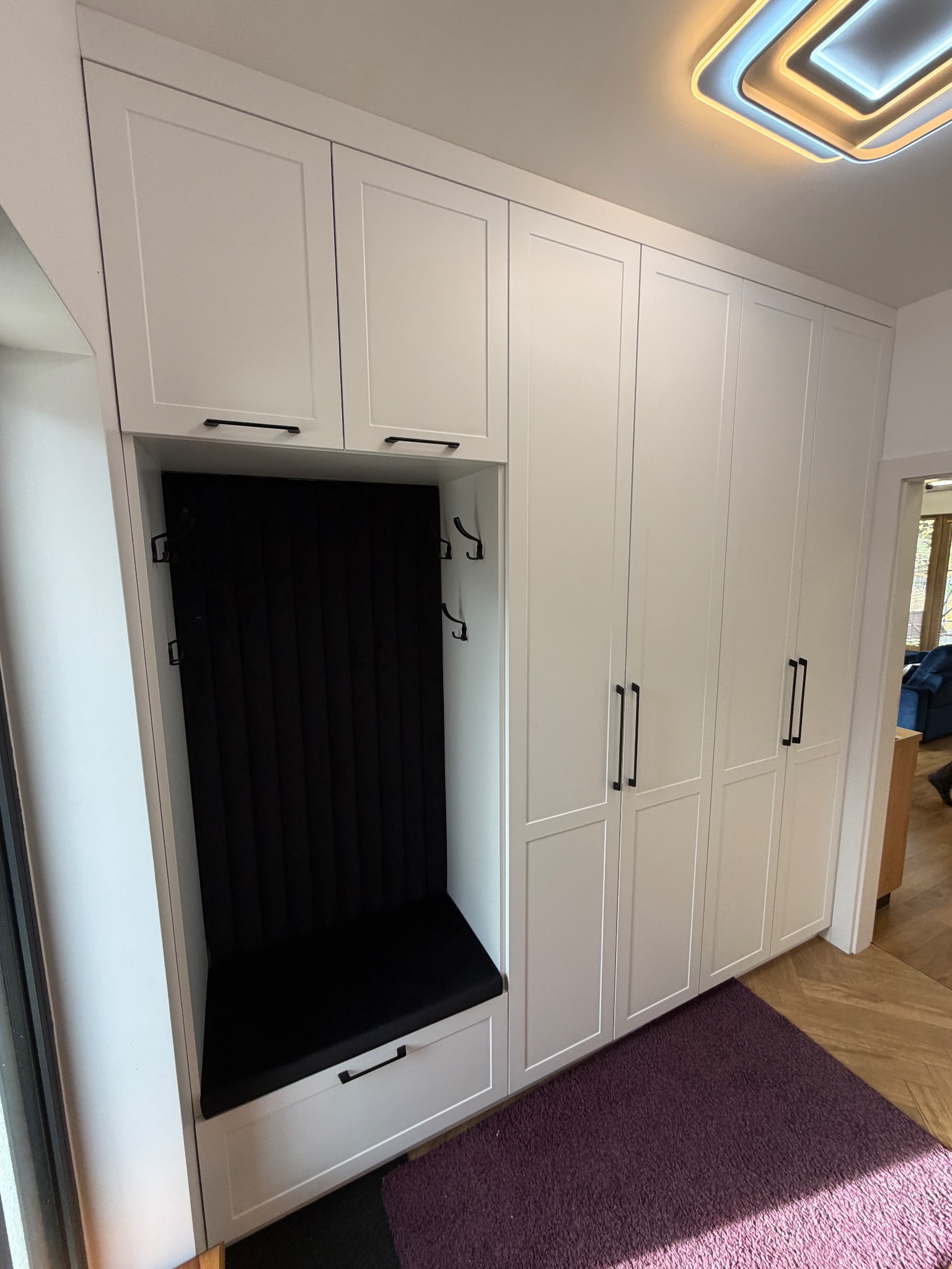 Biała szafa wnękowa z siedziskiem i wieszakami, czarne uchwyty, fioletowy dywanik. Nowoczesny design, oświetlenie sufitowe. Funkcjonalne rozwiązanie do przedpokoju.