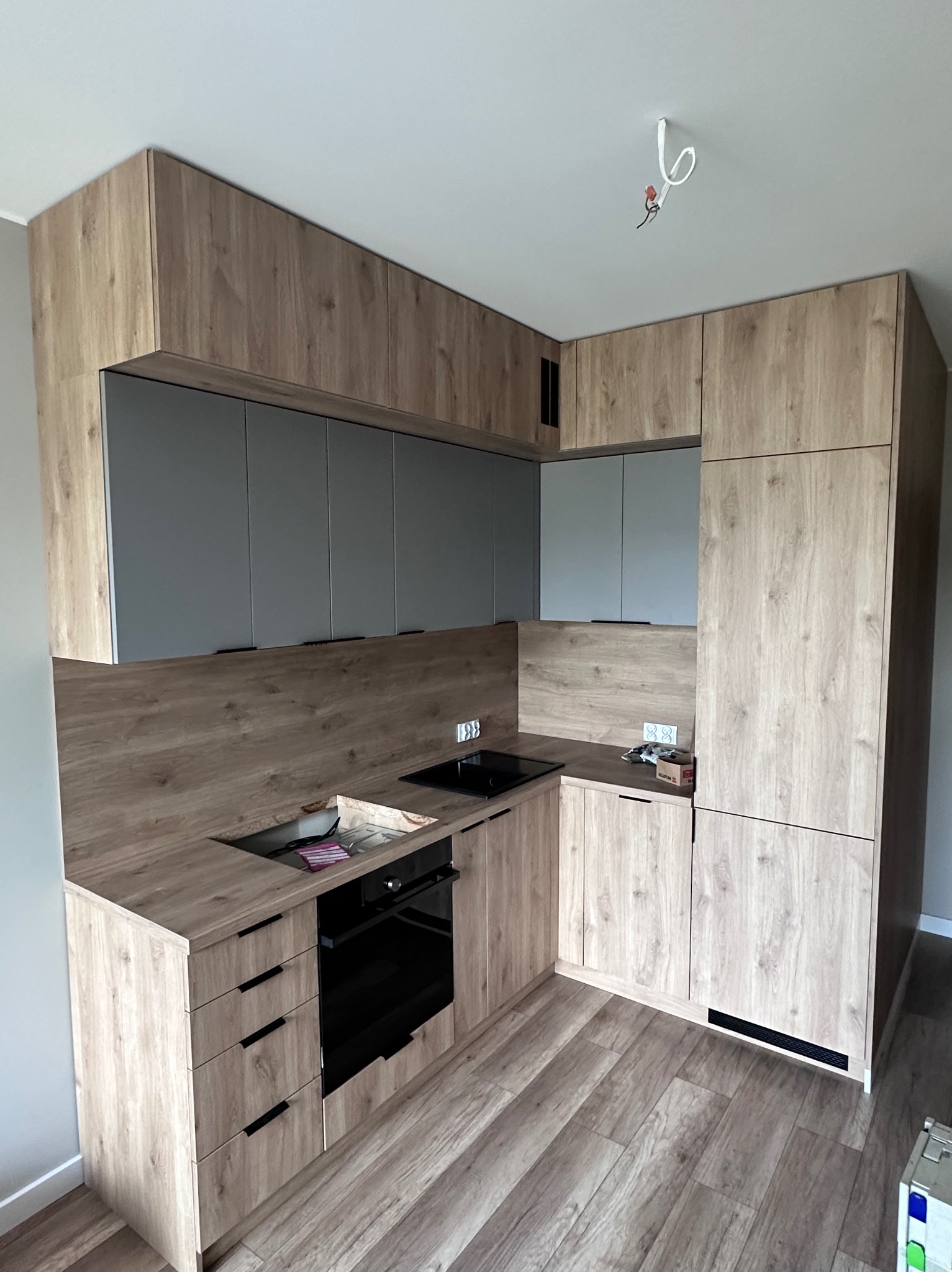 Nowoczesna kuchnia z drewnianymi frontami i szarymi szafkami wiszącymi. Uwagę zwraca minimalistyczny design i oświetlenie podszafkowe. Widoczny fragment podłogi.