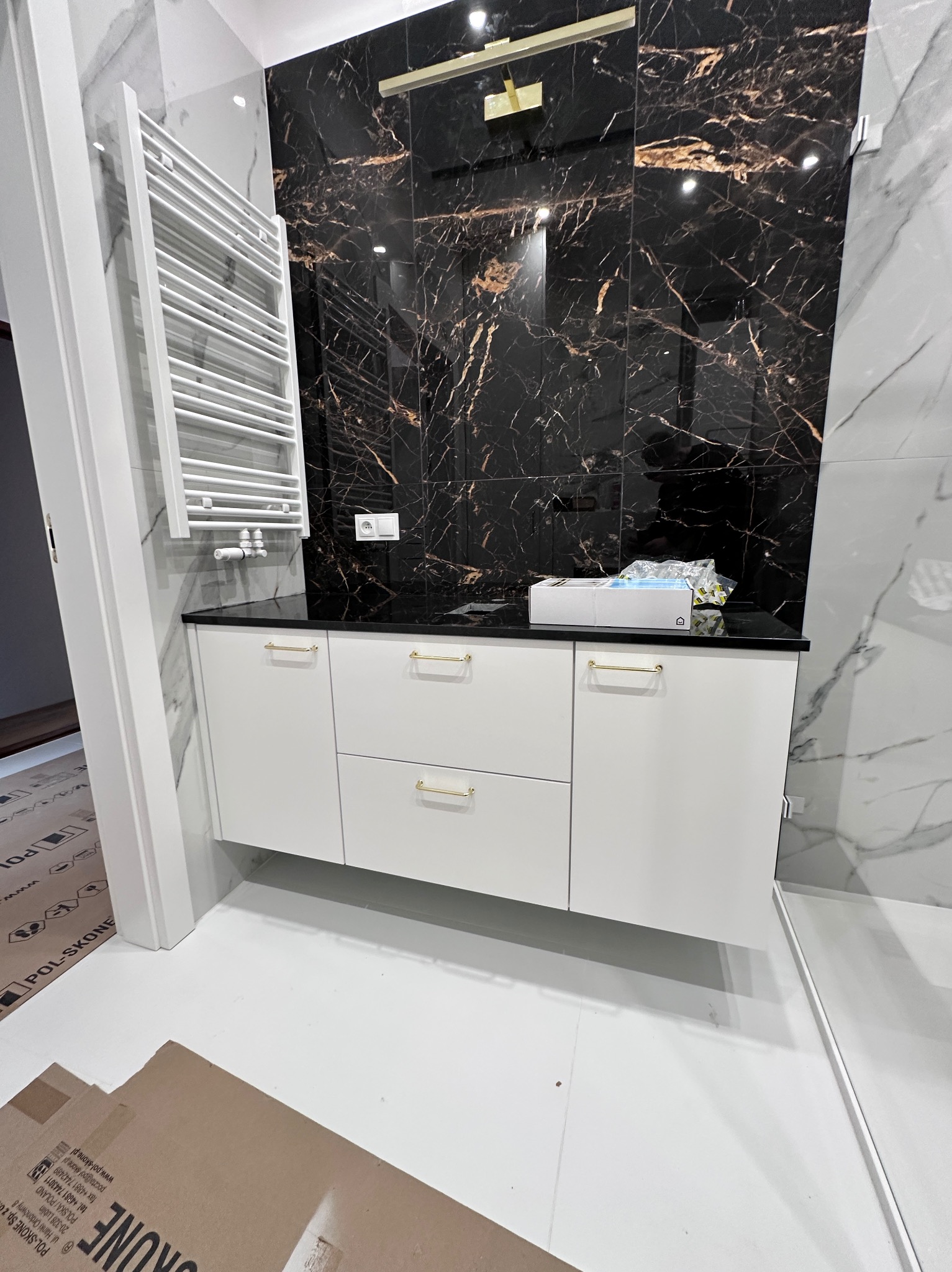 Nowoczesna łazienka z białą szafką pod umywalkę z złotymi uchwytami, czarnym blatem i marmurową ścianą. Widoczny grzejnik drabinkowy i fragment podłogi z kartonami.