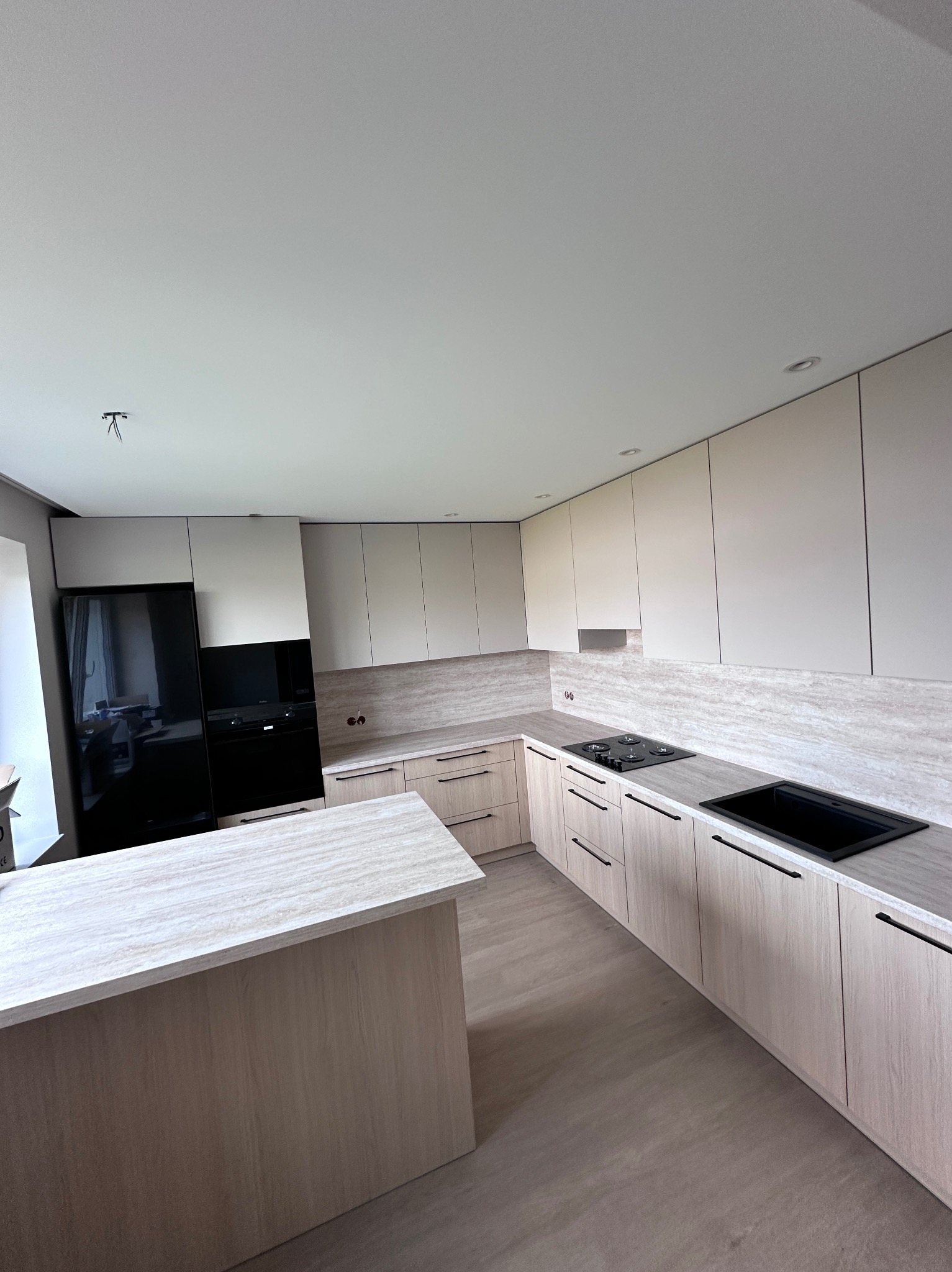 Jasna kuchnia na wymiar z wyspą, fronty w odcieniach beżu i szarości, czarne akcenty AGD i armatury. Minimalistyczny design, oświetlenie punktowe.