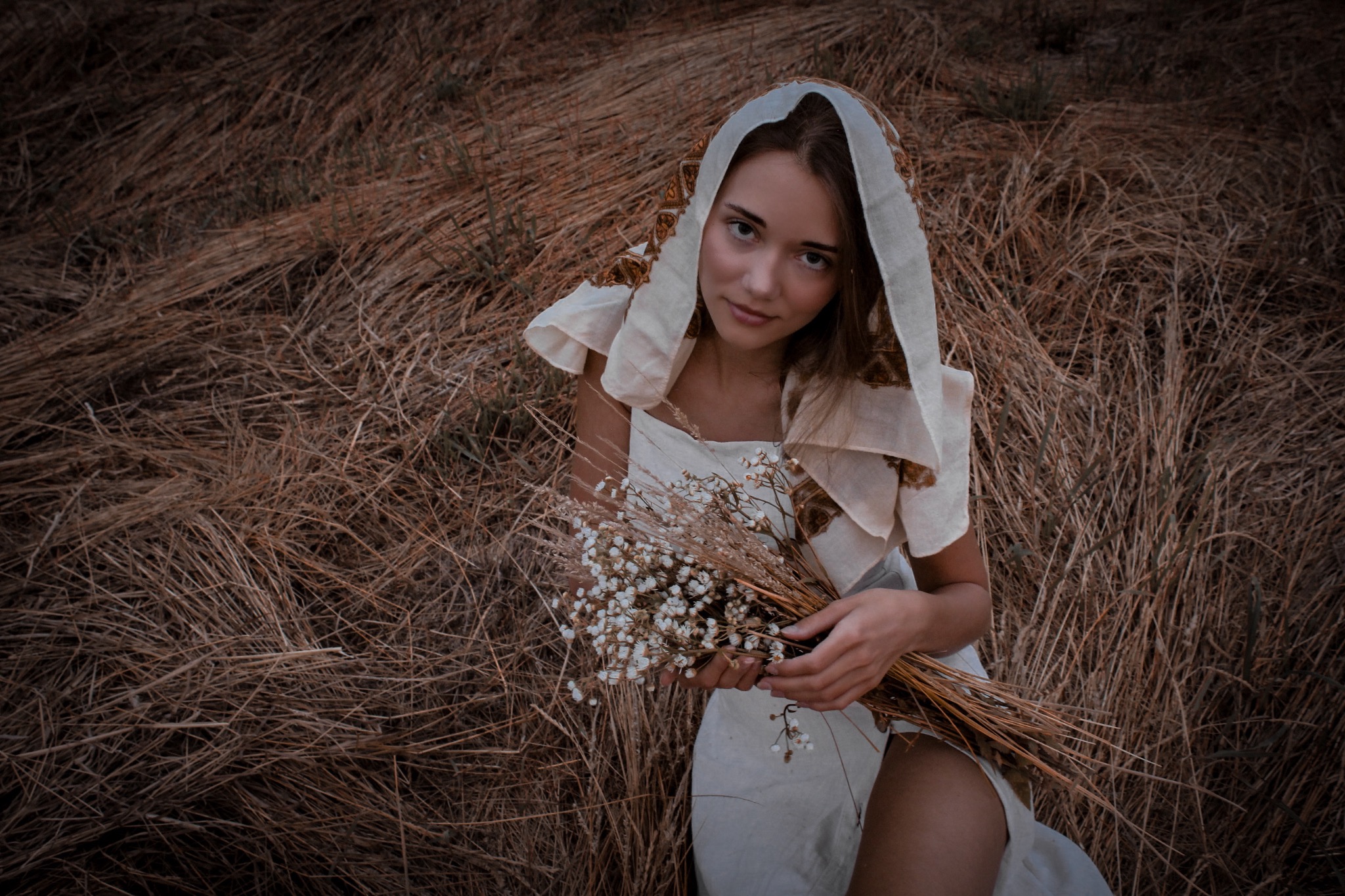 Młoda kobieta w jasnej sukience i chuście na głowie siedzi na wyschniętej trawie, trzymając bukiet polnych kwiatów.