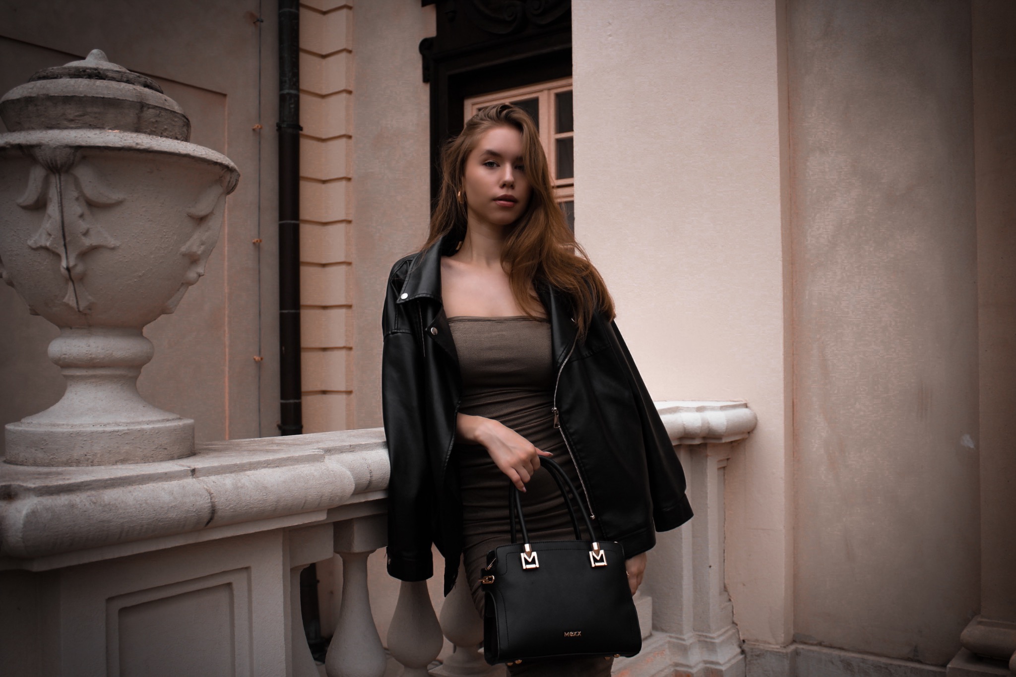 Portret młodej kobiety w skórzanej kurtce i dopasowanej sukience, trzymającej czarną torebkę na tle klasycznej architektury z kamienną balustradą i dekoracyjną urną.