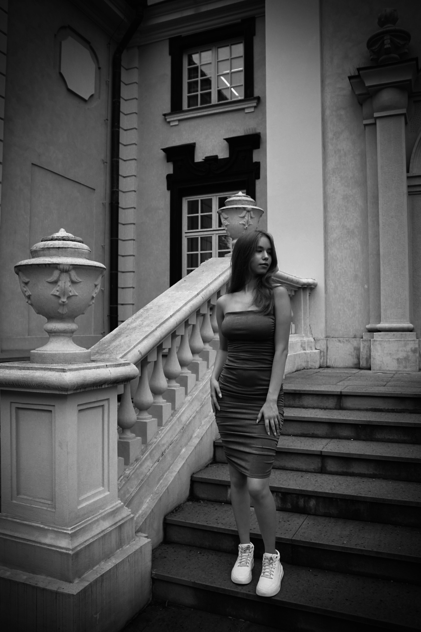 Młoda kobieta w dopasowanej sukience i sportowych butach pozuje na schodach kamienicy z klasyczną balustradą i urnami, fotografia czarno-biała.