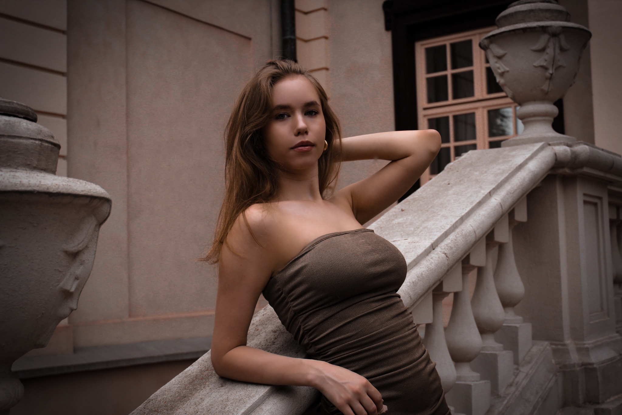 Portret młodej kobiety w brązowej sukience bez ramiączek, opierającej się o kamienną balustradę z dekoracyjnymi urnami w tle.
