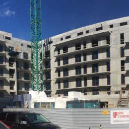 PROJEKTOWANIE KONSTRUKCJI BUDOWLANO-INŻYNIERYJNYCH TOMASZ SZUMSKI - Budynek mieszkalny w stanie surowym, z widocznym żurawiem budowlanym i fragmentem ogrodzenia z logotypem wykonawcy.