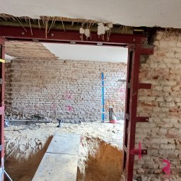 PROJEKTOWANIE KONSTRUKCJI BUDOWLANO-INŻYNIERYJNYCH TOMASZ SZUMSKI - Wnętrze budynku w trakcie wzmacniania konstrukcji ściany ceglanej, widoczne stalowe profile i wykop w podłodze.