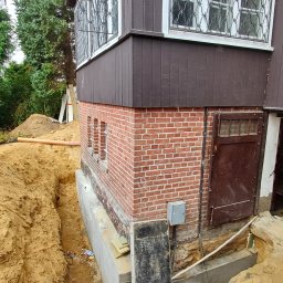 PROJEKTOWANIE KONSTRUKCJI BUDOWLANO-INŻYNIERYJNYCH TOMASZ SZUMSKI - Budynek z ceglaną podmurówką i drewnianą elewacją w trakcie prac ziemnych, wykop wzdłuż fundamentów, widoczne rury i elementy konstrukcyjne.