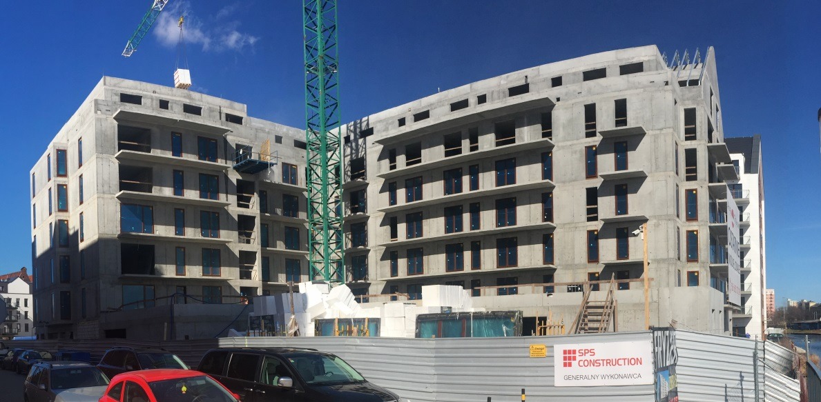 Budynek mieszkalny w stanie surowym, z widocznym żurawiem budowlanym i fragmentem ogrodzenia z logotypem wykonawcy.