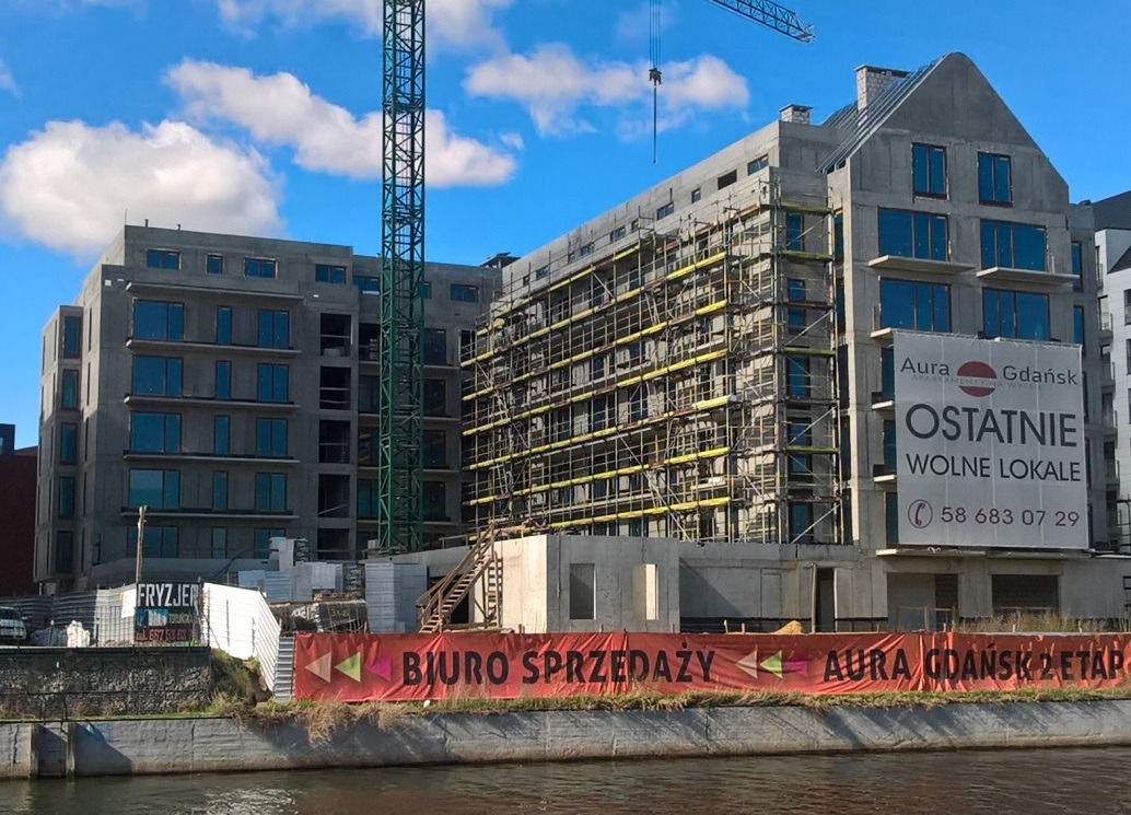 Wielopiętrowy budynek w stanie surowym, z rusztowaniami i dźwigiem, widoczny baner reklamowy z napisem 'Ostatnie wolne lokale' i 'Biuro sprzedaży'.