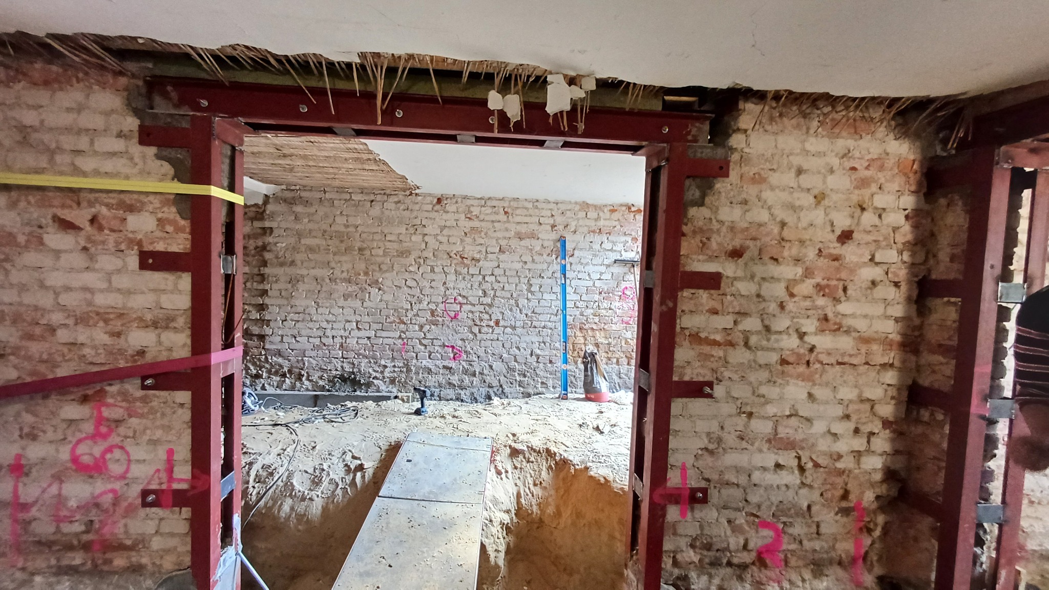 Wnętrze budynku w trakcie wzmacniania konstrukcji ściany ceglanej, widoczne stalowe profile i wykop w podłodze.