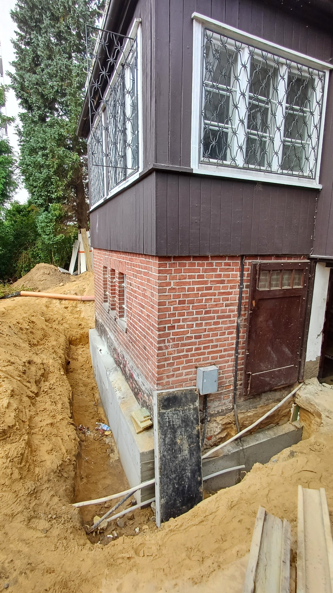 Budynek z ceglaną podmurówką i drewnianą elewacją w trakcie prac ziemnych, wykop wzdłuż fundamentów, widoczne rury i elementy konstrukcyjne.